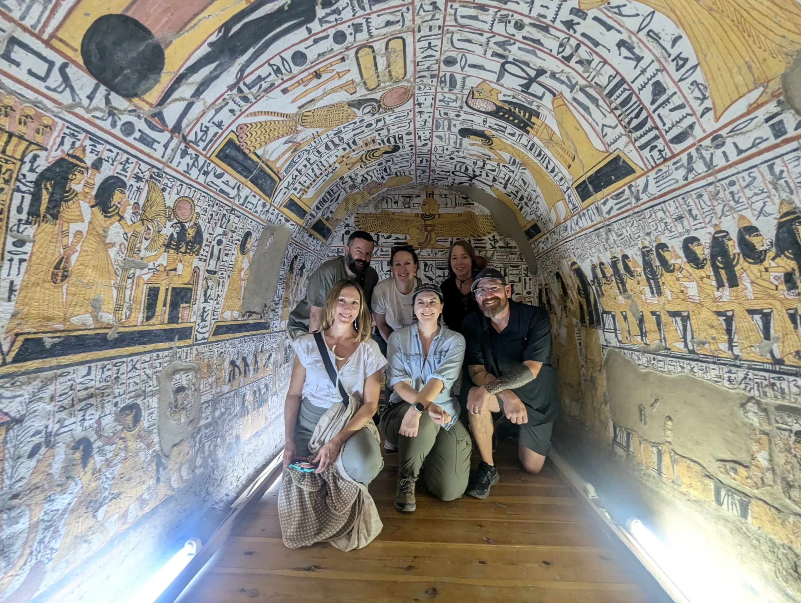 A group of 6 people smile for a group photo inside a tomb with intricate yellow black white and red paintings at Deir el medina