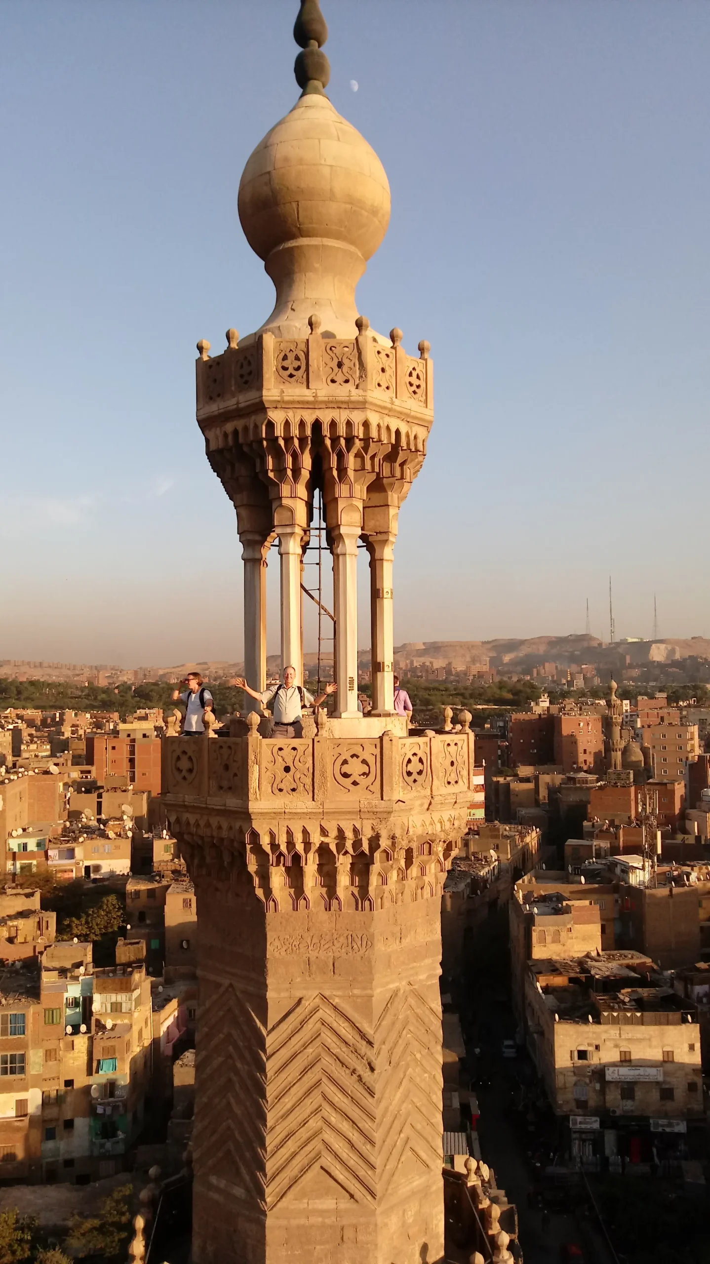Bab Zuweila minaret in the golden hour. A man stands on the overlook of the minaret looking happy and raising his arms. the smaller buildings of Cairo stretch in all directions to the Horizon.