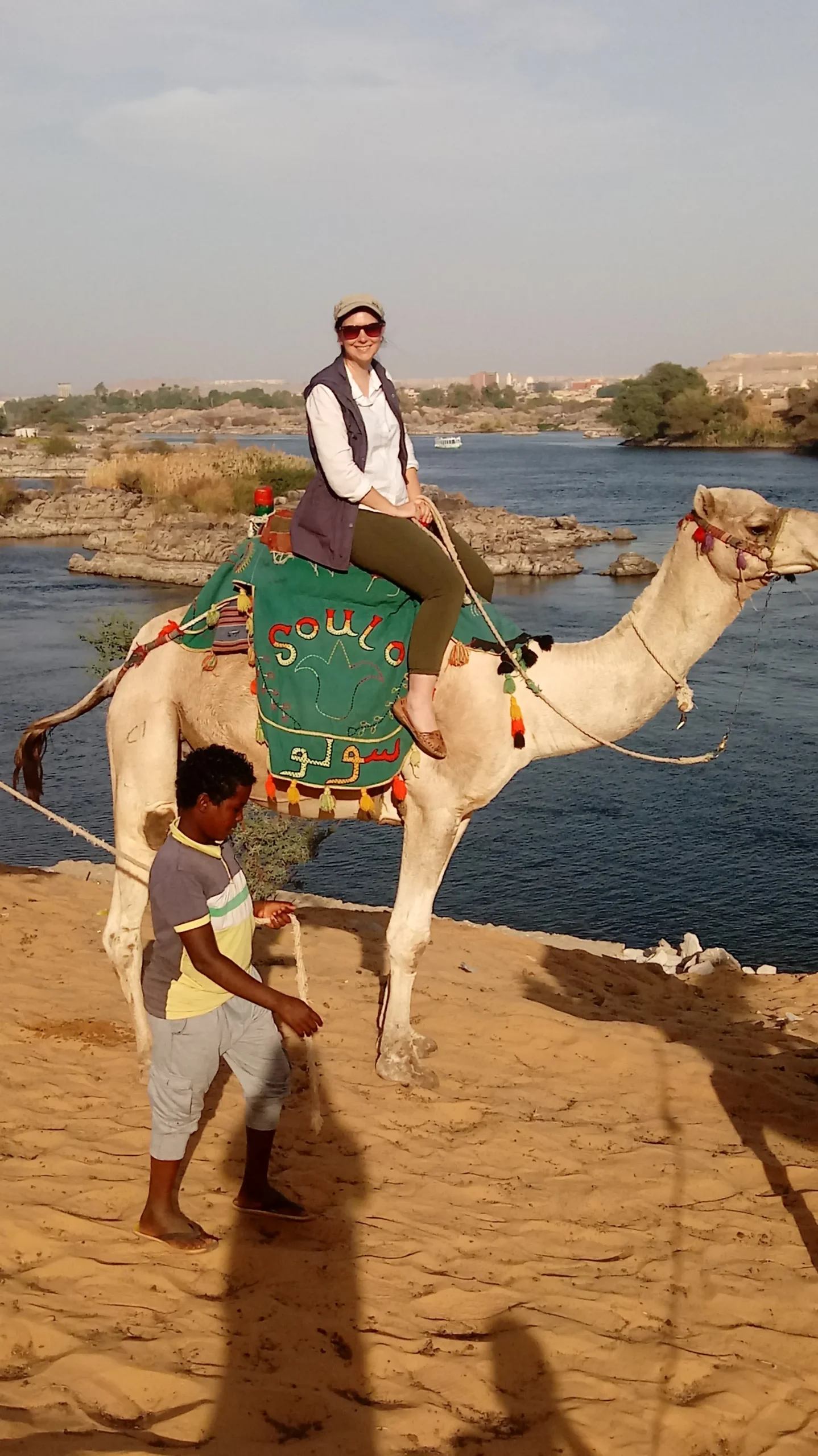 Beth on a camel, on a cliff above the Nile in Aswan. the camel is being led by a Nubian boy. the saddle on the camel is green and colorful and says "soulo" in Arabic and English.