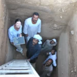 Five people are in a deep excavation pit looking up at the camera