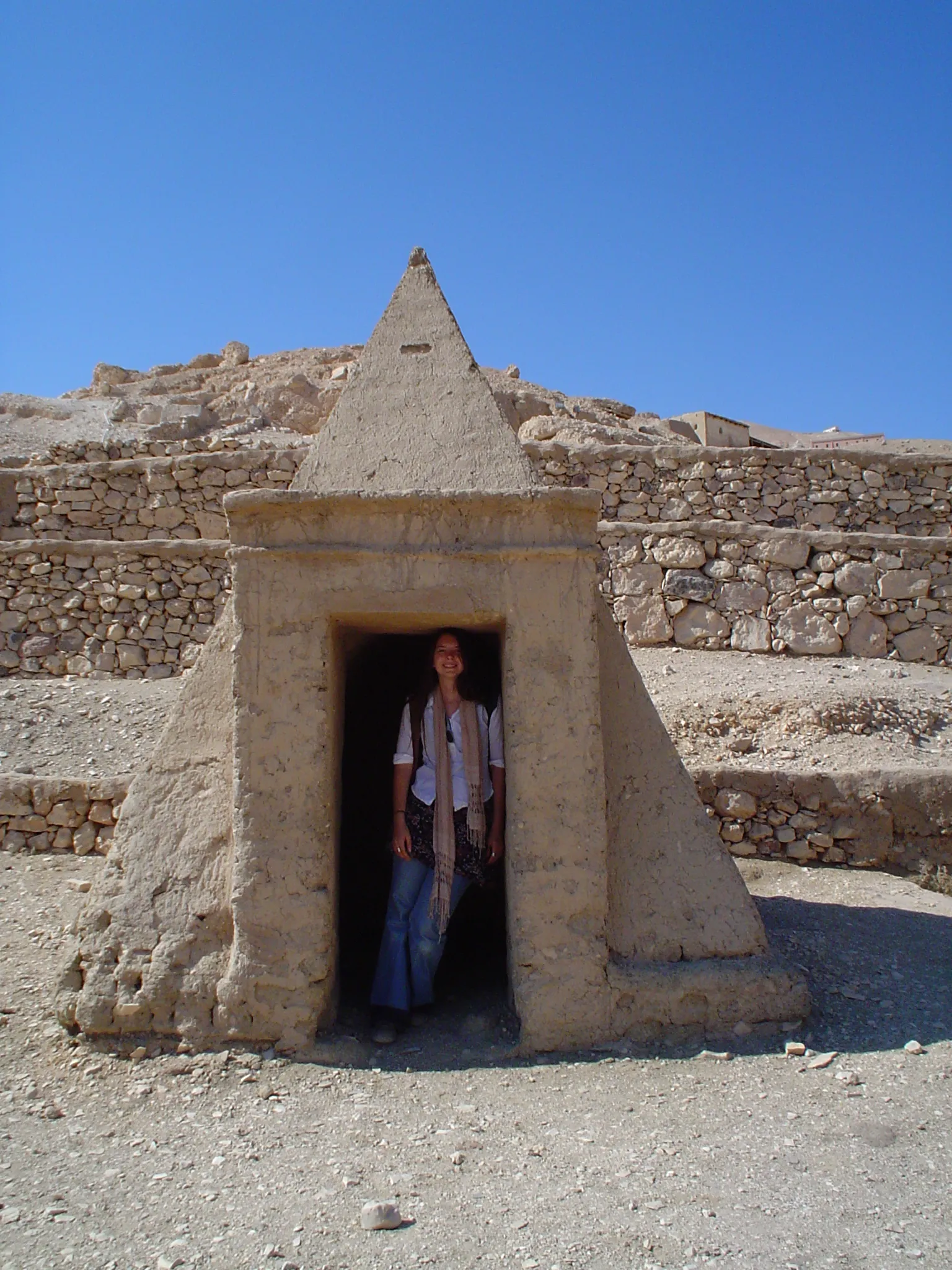 A small, steep-sides pyramid that is just larger than the doorway entrance carved into it. Beth stands in the doorway looking out. At Deir el Medina.