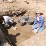Two people in a shallow square excavation pit, carefully sweeping up dirt and putting it in plastic black baskets