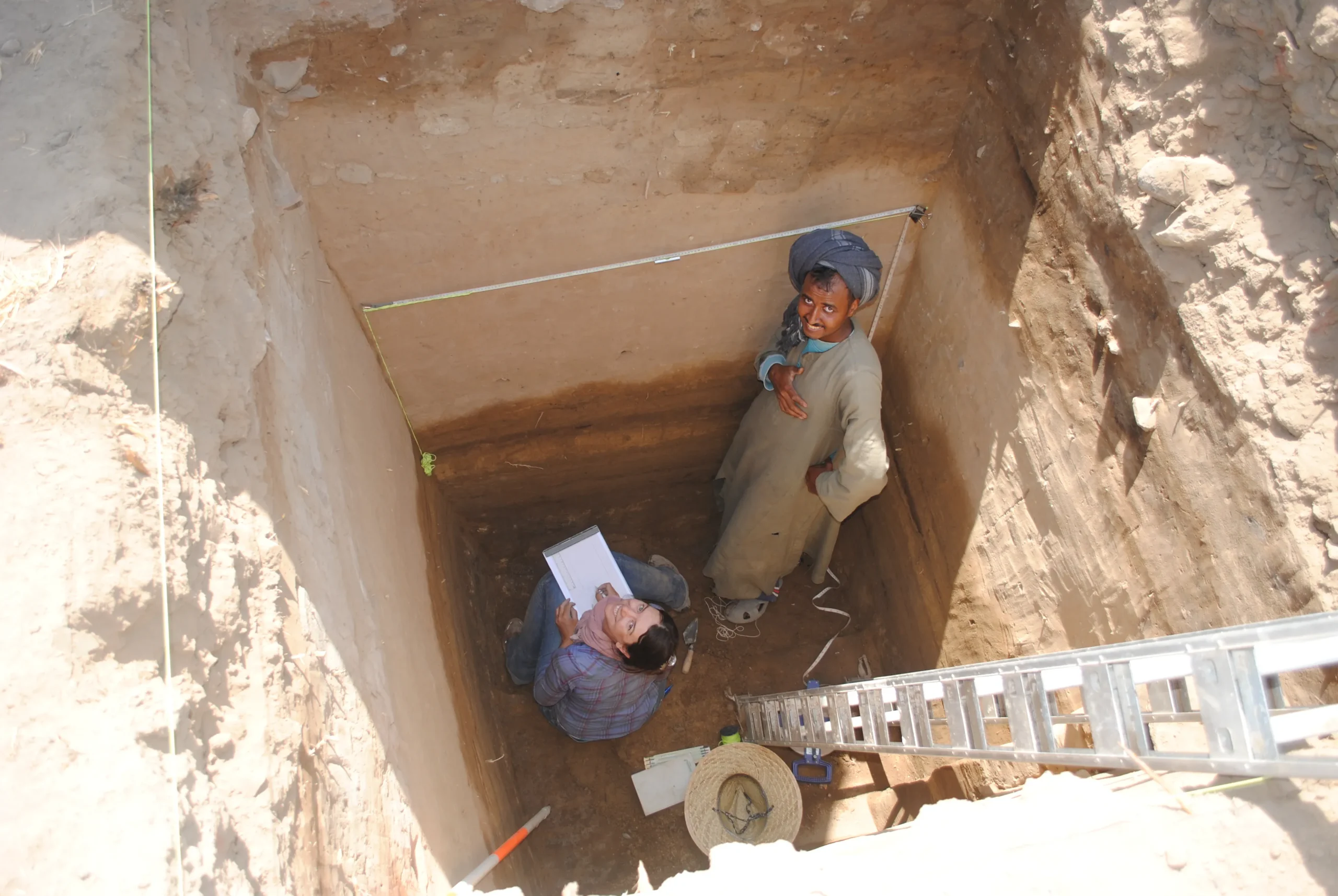 Teo people inside a deep square test pit. An Egyptian man in traditional head wrap and galabiya stands, and Beth sits with a notepad in front of her, both are looking up at the camera and smiling.