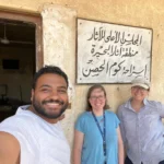 THree peopel stand in front of a sign in Arabic. they are dirty but smiling.
