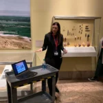 Beth stands in a black suite in front of a wall display of stone tools and gestures at a computer on a cart