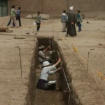 two peopel sit in a long narrow trench with measuring tapes making drawings. Egyptian villagers in local dress walk nearby