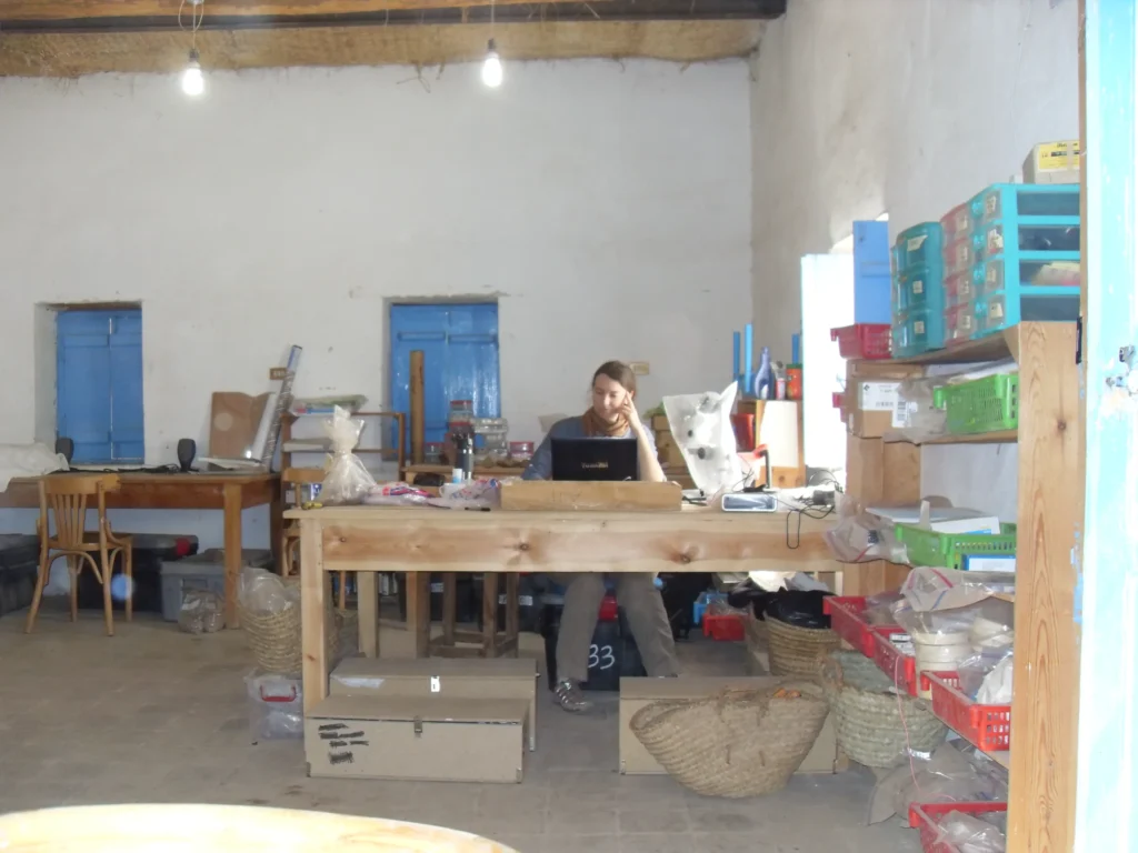 The image shows Beth at a wooden desk in a cluttered room. She is looking at a computer with her hand to her forehead deeply focused on and doing archaeological work, probably in a similar pose as anyone who might be focusing to read terms and conditions.