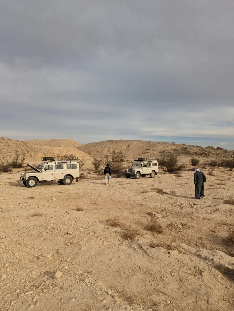 Day in the Life of an Archaeologist in Egypt The image shows a tan desert landscape with a few low hills, scrubby bushes, and a cloudy sky. There are two off-white landrovers one with the hood propped open and a man in a galabiya is walking near the vehicles. The image shows the setting of the desert archaeology.