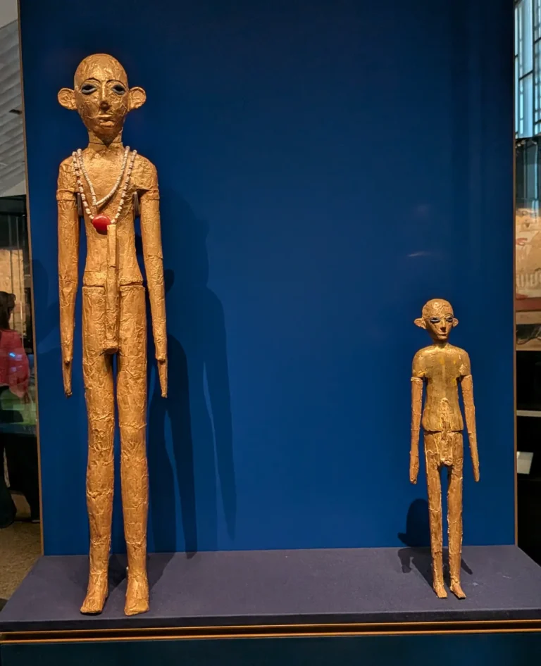 Two golden Statuettes, both depicting men with large ears, eyes inlaid with Lapis Lazuli, and large penis sheaths. One is twice the size of the other and wears a necklace of shell and carnelian.