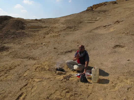 A man sits cross-legged on a sandy desert hill next to a small, smoking campfire. Various supplies, a large plastic container, and a dark bag rest on the sand beside him.