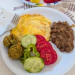 Egyptian Cuisine: 7 surprising things for first-time travelers A white plate serves a traditional Egyptian breakfast with a round egg omelet, a serving of brown fava beans (ful), green taameya (falafel) pieces, and sliced tomatoes, cucumbers, and peppers.