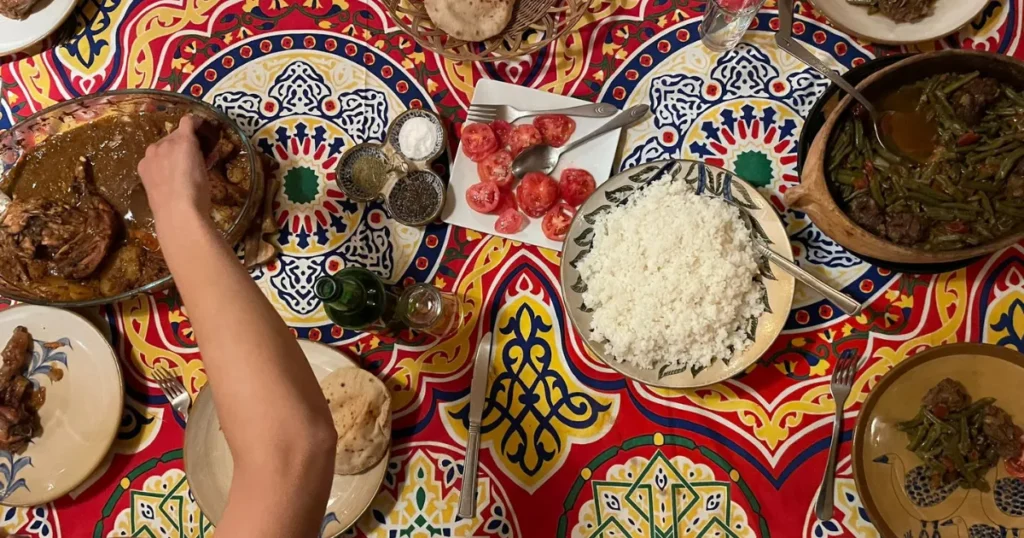 Overhead view of a meal spread on a colorful patterned tablecloth. The meal includes a large plate of white rice, sliced tomatoes, green bean stew, saucy chicken, and pita bread.