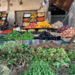 Egyptian Cuisine: 7 surprising things for first-time travelers A vibrant outdoor Egyptian market stall overflows with piles of fresh green beans, leafy herbs, cucumbers, eggplants, onions, and stacked crates of oranges, apples, and bananas.