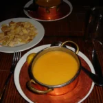 Egyptian Cuisine: 7 surprising things for first-time travelers A warm orange lentil soup sits in a copper bowl with brass handles on a dark table, accompanied by a small side plate of crispy, cubed pita bread pieces and a lemon wedge