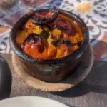 Egyptian Cuisine: 7 surprising things for first-time travelers A small clay pot sits on a woven mat, holding a bubbling Egyptian vegetable tagine with charred tomato slices on top, placed over a colorful geometric patterned table.