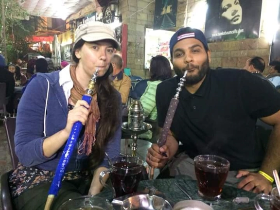 A man and woman are each smoking shisha at a lively cafe. Two mugs of dark tea sit on the table, surrounded by patrons in a bustling, atmosphere.