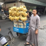 Egyptian Cuisine: 7 surprising things for first-time travelers A vendor in a grey robe stands next to a blue tricycle cart loaded with bunches of yellow bananas on a city street.
