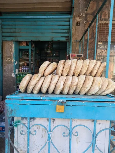 Two rows of small thick, golden-brown bread loaves stand upright in on top of a blue metal cart positioned outside a shop entrance.