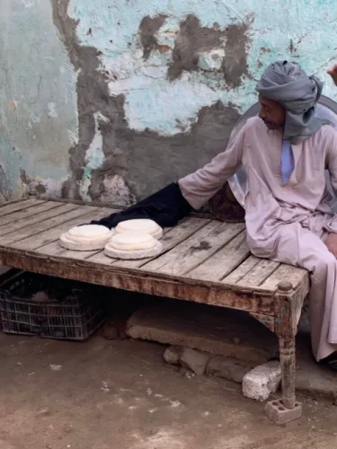 A man in a traditional robe and headscarf sits on a wooden slatted bench next to three round, unbaked dough rounds resting on circular mats against a weathered wall.