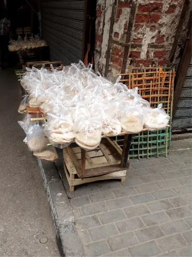 Dozens of transparent plastic bags filled with round pita bread sit on a wooden frame on a street corner in front of a textured brick wall