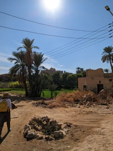 The image shows the walk back through the village, there is a dirt road with a small mud brick building on one side, and a a rock lined firepit. Behind the road and house is a lush green agricultural field with palm trees at the edge.