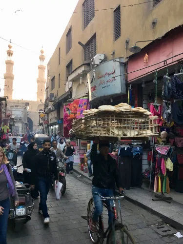 A cyclist carrying a large multi-tiered tray of bread maneuvers through a crowded market alley lined with shops, a tall stone minaret visible in the background, as pedestrians walk past.