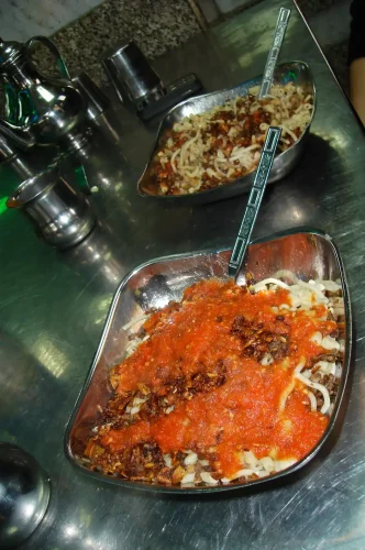 Two metal bowls of Egyptian koshari sit on a stainless steel table, showing layers of rice, macaroni, lentils, and crispy onions topped with a bright red tomato sauce.