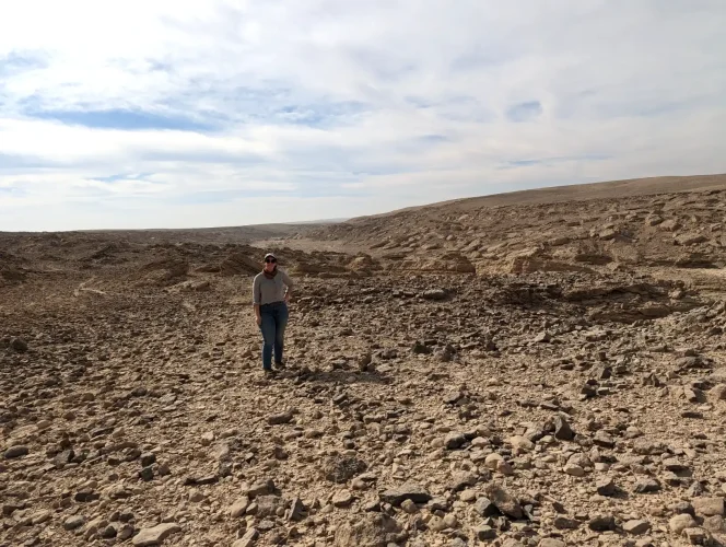 Beth is in a vast flat desert landscape with lightly rolling hils in the distance, a bright blue sky with wispy clouds and a gravelly surface.