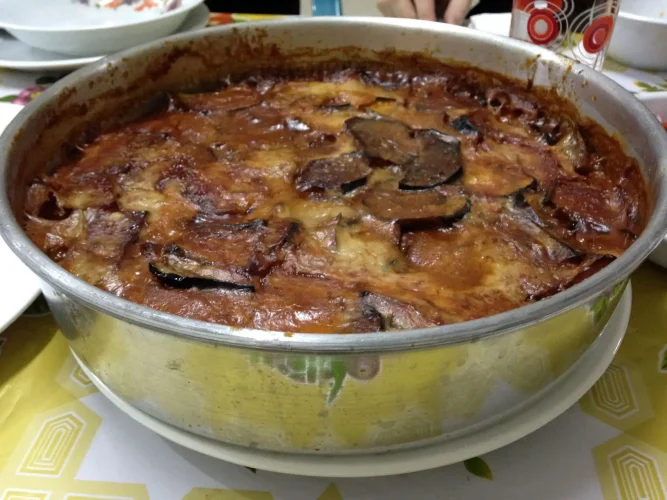 A large round metal pan holds a freshly baked Egyptian moussaka, featuring layers of sliced eggplant glistening with oil and baked into a thick tomato sauce.