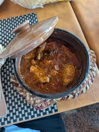 An open clay tagine dish reveals a bubbling, thick red tomato stew with tender chunks of chicken, resting on a woven straw mat atop a restaurant table.