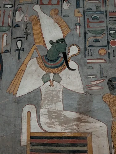 A painted image of the God Osiris. He is seated on a low chair, his body is wrapped in white cloth like a mummy. his hands are exposed and hold a scepter and a flail. He wears a broad collar. the skin of his face and neck is green and he has a thin black stylized beard. Hea wears a conical white crown with golden feathers on either side of it. THe wall is a dark blueish gray and colorful hieroglyphs surround him.