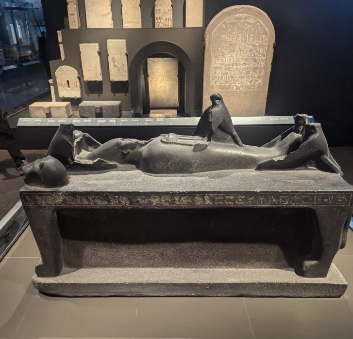 A basalt statue of the god Osiris laying on a funerary bed. the Goddess Isis in the form of a bird sits atop him.