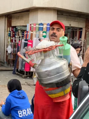 On the street in front of a textile shop an Egyptian street vendor wearing a red cap carries a large, traditional metal tea vessel strapped to his chest. Empty glasses are secured to the outside of the vessel.