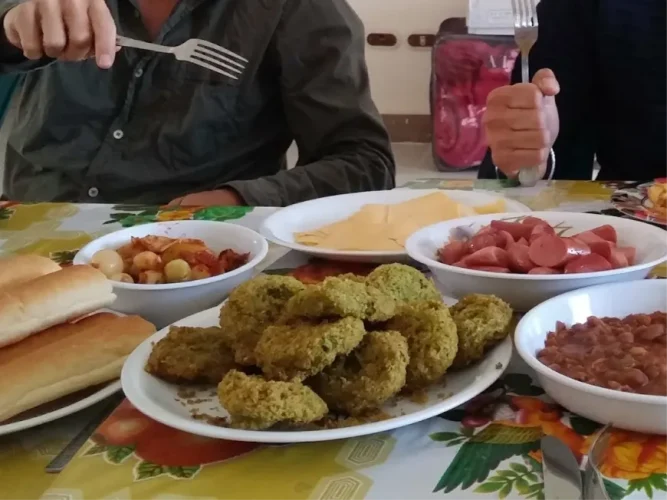 A close-up of a breakfast spread features a large central plate piled high with crispy, golden-green taameya (Egyptian falafel), surrounded by bowls of sliced sausages, fava beans, and fresh bread.
