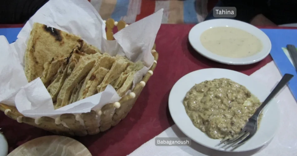 A plate of creamy tahina dip sits next to one of textured babaganoush dip and a woven basket with several pieces of cut balady pits bread on a maroon tablecloth.