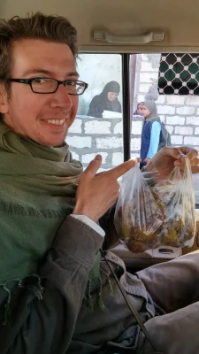 A man with glasses and a green scarf smiles inside a vehicle, holding up a plastic bag loaded with round balls of taameya. Outside the window, 2 local women stand near a white brick wall.