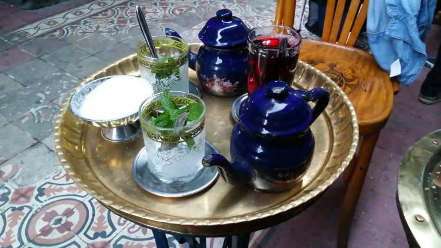A round brass tray holds two dark blue teapots, two glasses filled with fresh mint leaves, a glass of dark red tea, and a small metal bowl filled with white sugar.
