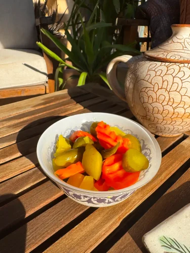 A white patterned bowl filled with colorful pickled vegetables, including green pickles and crinkle-cut orange carrots, sits on a wooden slatted table next to a patterned ceramic teapot