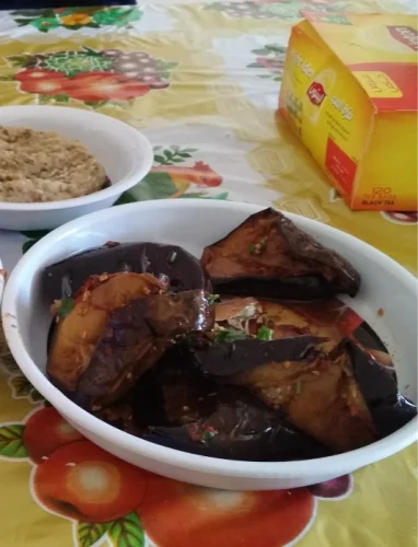 A white oval bowl filled with glossy, marinated Egyptian pickled eggplant slices sits on a patterned tablecloth next to a small side dish and a yellow Lipton tea box.
